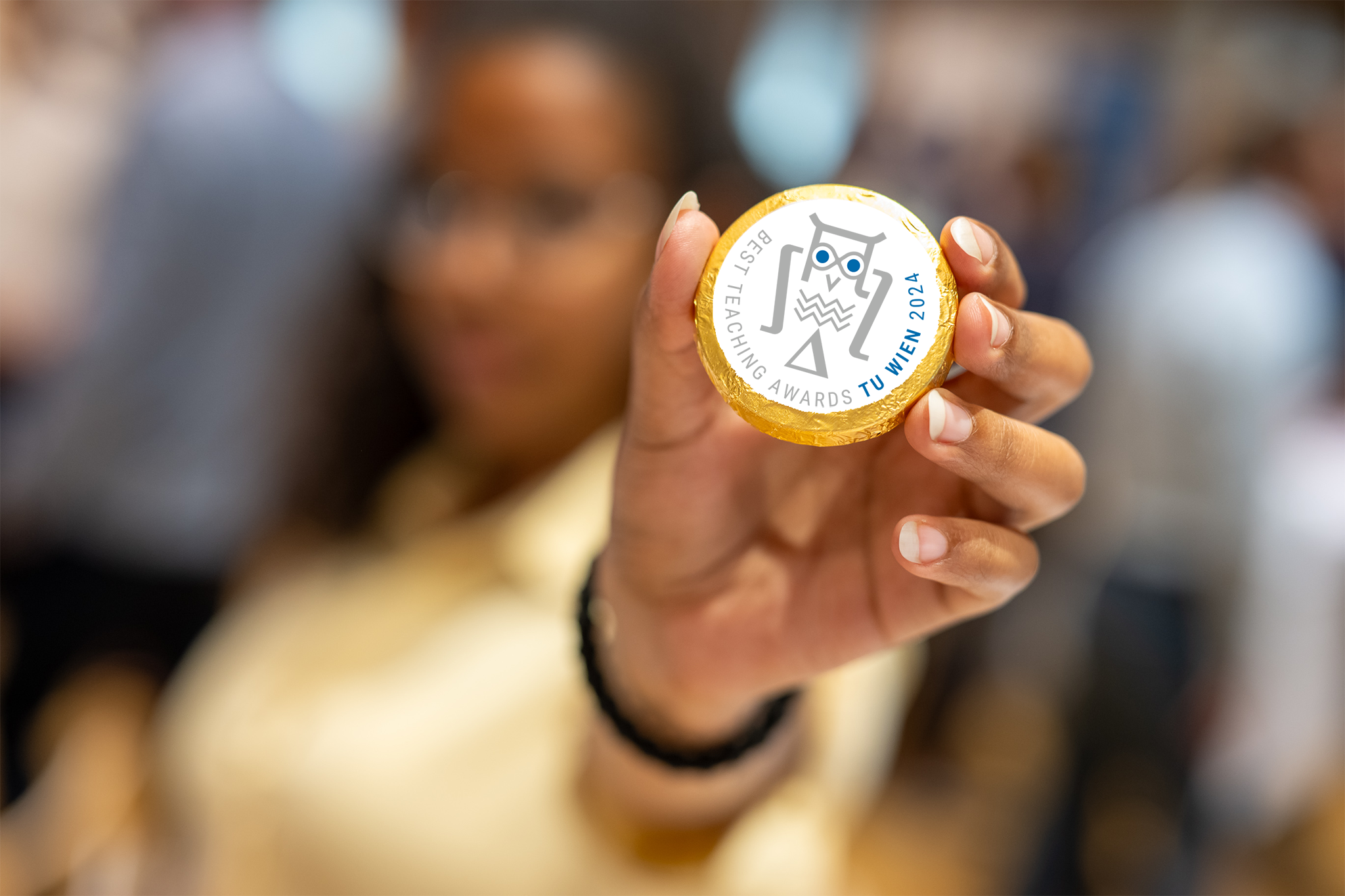 Logo Best Teaching Award auf Schokotaler und Frau verschwommen in Hintergrund, die Schokotale in der Hand hält und in die Kamera streckt