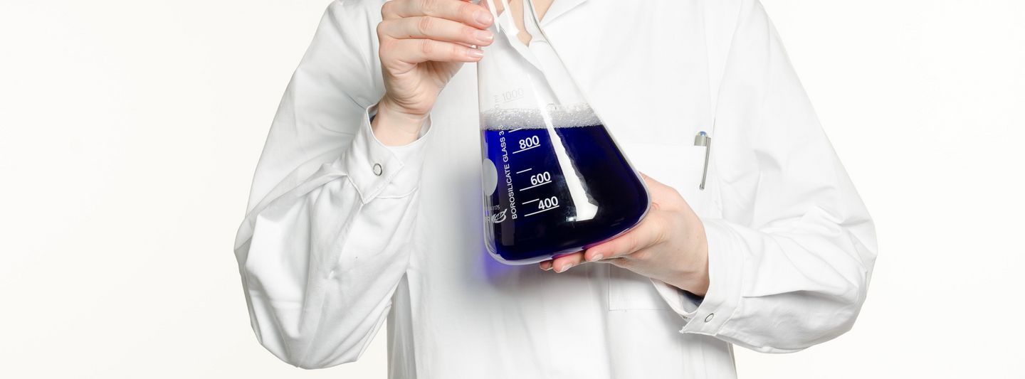 a person in a white coat holds an Erlenmeyer flask filled with a blue liquid