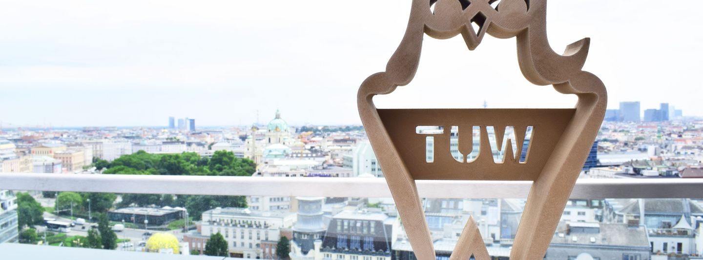 Im Vordergrund ist die Trophäe der "Best Teaching Awards" zu sehen. Im Hintergrund ist ein Panorama des Karlsplatzes zu sehen.