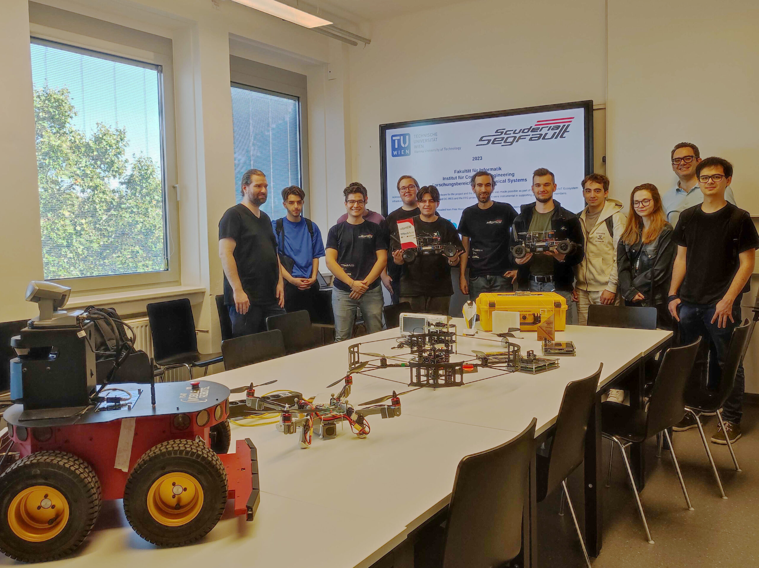 Students holding two racing cars, standing behind a table with several robots