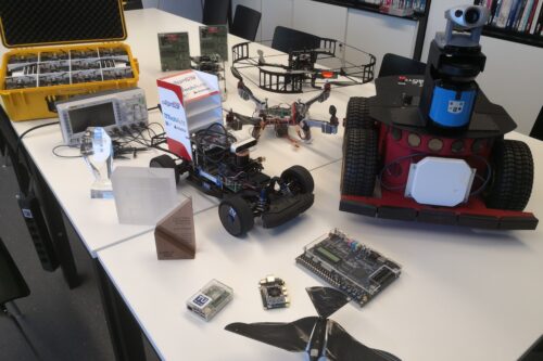 racing car, rover, bird-shaped robot, some quadcopters, and several computing platforms on a table