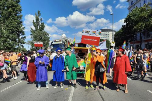 Regenbogenparade Menschen, im Vordergrund 6 Personen mit bunten Hochschultalaren